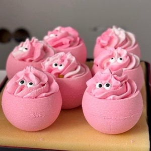 Op de afbeelding: Roze badbruisballen met een werveling van roze glazuur en kleine witte en zwarte snoepoogjes. De badbruisballen zijn gerangschikt op een gele en bruine tray. De badbruisballen zijn bolvormig en hebben een grillig uiterlijk.
