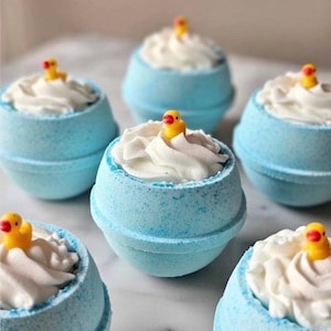 Puede incluir: Seis bombas de baño azules, cada una cubierta con jabón batido blanco y un pequeño patito de goma amarillo. Las bombas de baño tienen una superficie texturizada y están dispuestas sobre una superficie de mármol blanco.