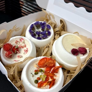 Puede incluir: Una caja de regalo blanca con una ventana transparente contiene cuatro pequeñas bombas de baño blancas y redondas. Cada bomba de baño está decorada con flores secas en tonos de rojo, naranja, morado y rosa. La caja está forrada con papel marrón.