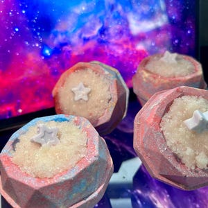 May include: Four star-topped bath bombs with a geometric design. The bath bombs are pink, blue, and white with a glittery finish. The background is a colorful nebula.