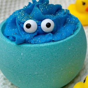 May include: A teal bath bomb shaped like a monster with blue frosting and googly eyes. The frosting is sprinkled with glitter. Two yellow rubber duckies are in the background.