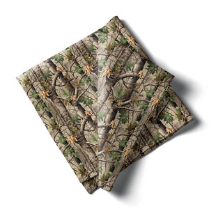 May include: A folded square fabric with a camouflage pattern. The design features a mix of brown, green, and tan colors, mimicking a forest environment with tree branches and leaves. The fabric is likely a handkerchief or a similar accessory.