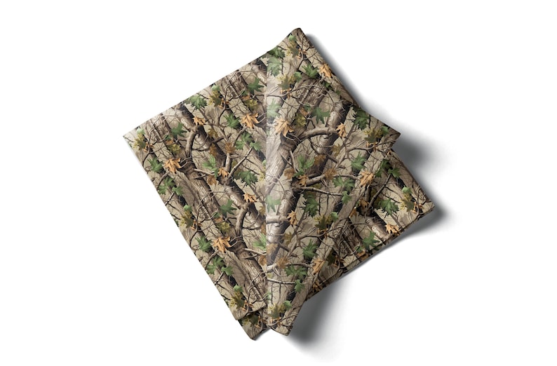 May include: A folded square fabric with a camouflage pattern. The design features a mix of brown, green, and tan colors, mimicking a forest environment with tree branches and leaves. The fabric is likely a handkerchief or a similar accessory.