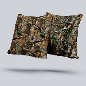 May include: Two square throw pillows with a camouflage pattern. The design features a mix of brown, tan, green, and gray, mimicking a forest environment with branches and leaves. The pillows appear to be made of a soft fabric.