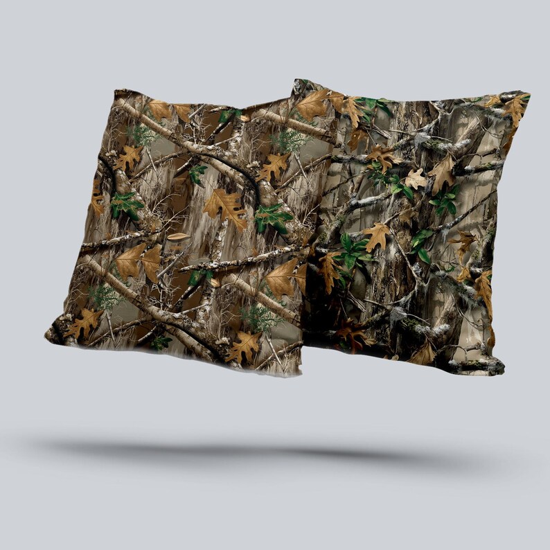 May include: Two square throw pillows with a camouflage pattern. The design features a mix of brown, tan, green, and gray, mimicking a forest environment with branches and leaves. The pillows appear to be made of a soft fabric.
