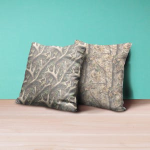 May include: Two square throw pillows with a camouflage pattern. One pillow features a pattern of deer antlers, while the other has a tree and leaf design. The pillows are in shades of brown, green, and gray, and rest on a wooden surface.