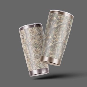 May include: Two stainless steel insulated tumblers with a camouflage pattern. The tumblers have a silver rim and base, and a lid. The camouflage design features shades of brown, green, and tan, resembling a forest scene.