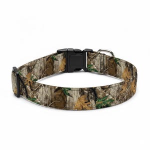 May include: A dog collar with a camouflage pattern in shades of brown, green, and tan. The collar has a black plastic buckle and a silver metal D-ring for leash attachment. The collar is designed for pets.