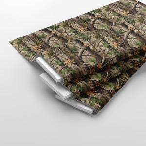 May include: A stack of camouflage fabric with a realistic forest pattern. The design features brown tree trunks, green leaves, and tan foliage. The fabric is folded over a white support structure.