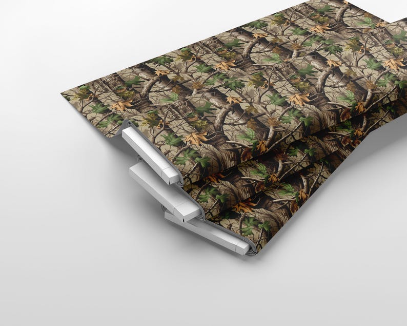 May include: A stack of camouflage fabric with a realistic forest pattern. The design features brown tree trunks, green leaves, and tan foliage. The fabric is folded over a white support structure.