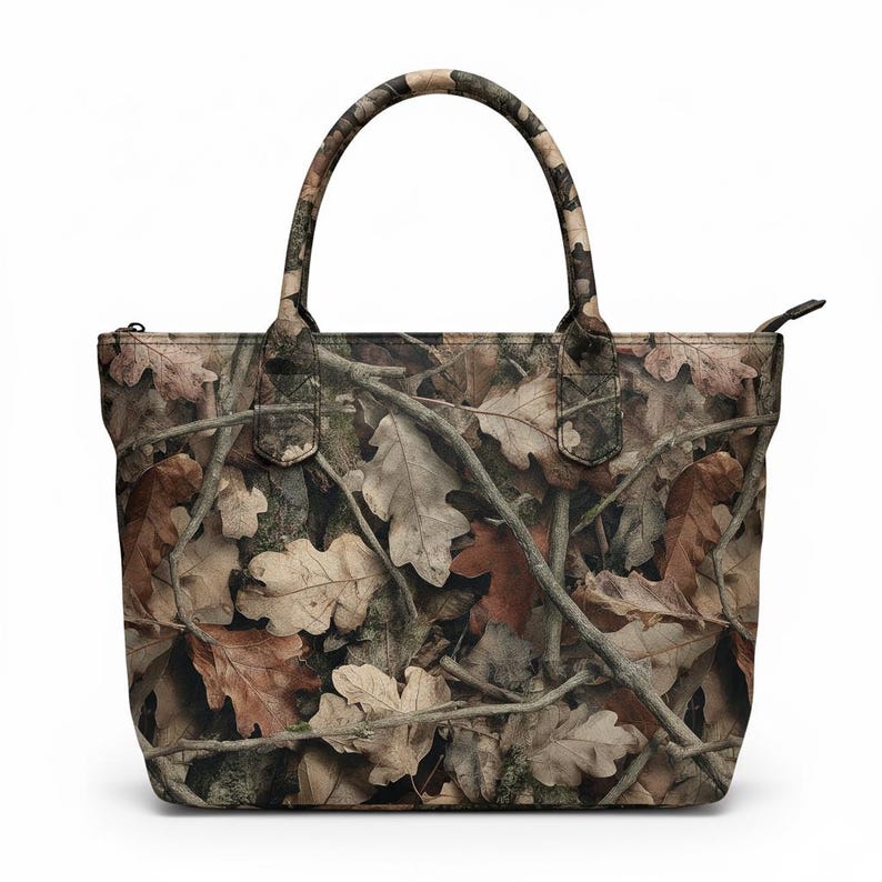 Puede incluir: Un bolso tote con un patr&oacute;n de camuflaje de hojas y ramas marrones y beige. El bolso tiene cierre de cremallera y dos asas. El dise&ntilde;o del bolso imita el suelo del bosque.