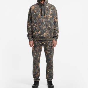 May include: A matching set of camouflage-patterned clothing, including a hooded sweatshirt and sweatpants. The set features a blend of brown, green, and tan hues, creating a woodland-inspired design. The sweatshirt has a front pocket and drawstring hood. The person is wearing black shoes.