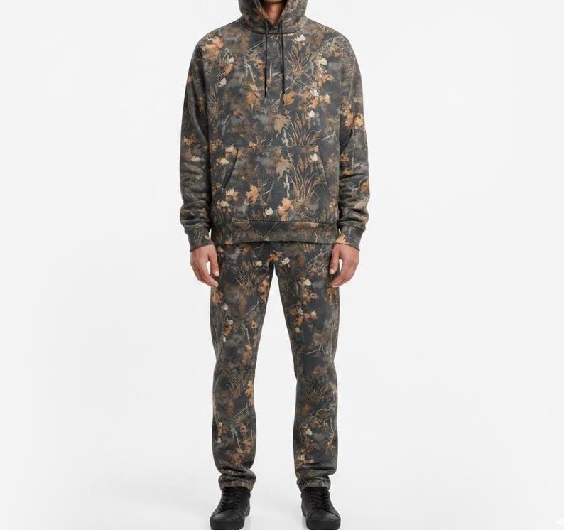 May include: A matching set of camouflage-patterned clothing, including a hooded sweatshirt and sweatpants. The set features a blend of brown, green, and tan hues, creating a woodland-inspired design. The sweatshirt has a front pocket and drawstring hood. The person is wearing black shoes.