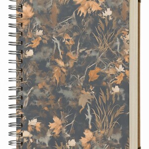 May include: Spiral-bound notebook with a camouflage pattern of autumn leaves in shades of brown, tan, and gray. The notebook has a dark gray background and metal corner protectors. The pages are cream-colored.