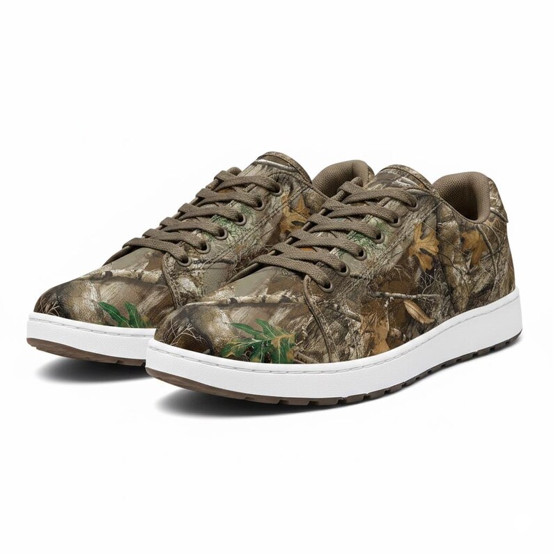 May include: A pair of low-top sneakers featuring a camouflage pattern in shades of brown, green, and tan. The shoes have white soles and brown laces. The design incorporates a realistic forest print.