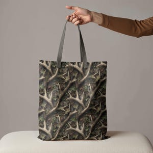 Puede incluir: Un bolso tote con un patr&oacute;n de camuflaje con cornamentas y hojas en tonos marrones, verdes y beige. El bolso tiene asas grises.
