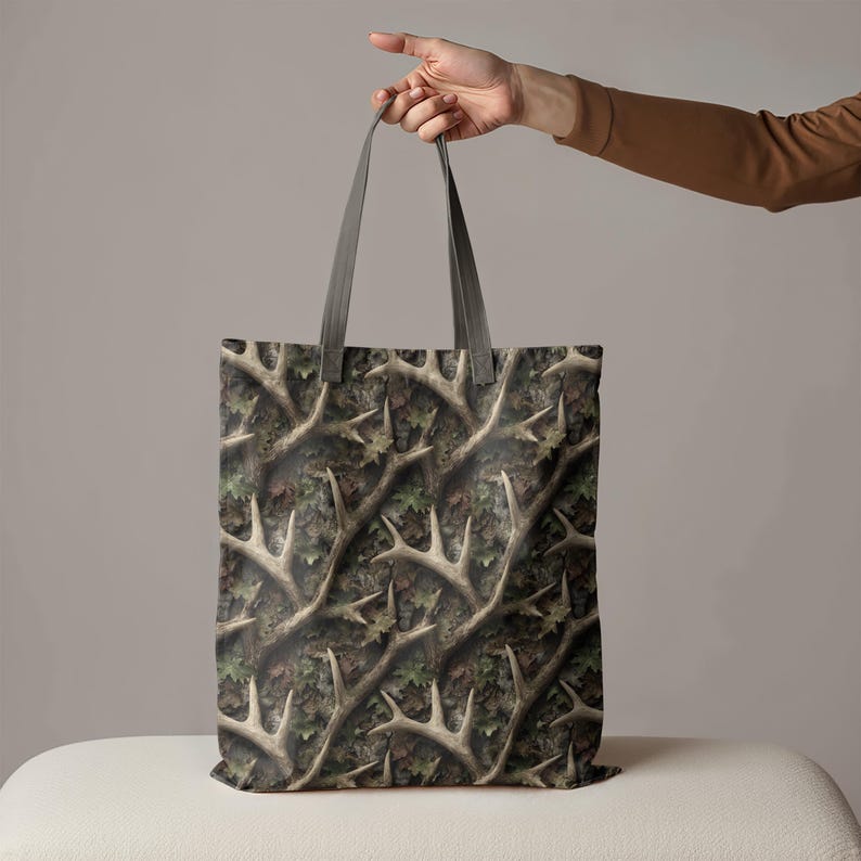 Puede incluir: Un bolso tote con un patr&oacute;n de camuflaje con cornamentas y hojas en tonos marrones, verdes y beige. El bolso tiene asas grises.