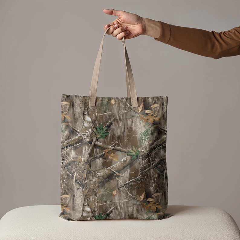 May include: A rectangular tote bag featuring a camouflage pattern in shades of brown, green, and tan. The bag has beige-colored handles and is held up by a person's hand. The bag's design mimics a forest environment.