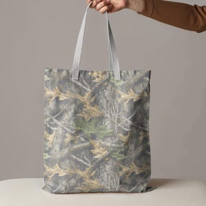 Puede incluir: Un bolso de mano con un estampado de camuflaje que presenta corteza de &aacute;rbol y hojas en tonos marrones, verdes y grises. El bolso tiene asas de color gris claro y lo sostiene una mano.
