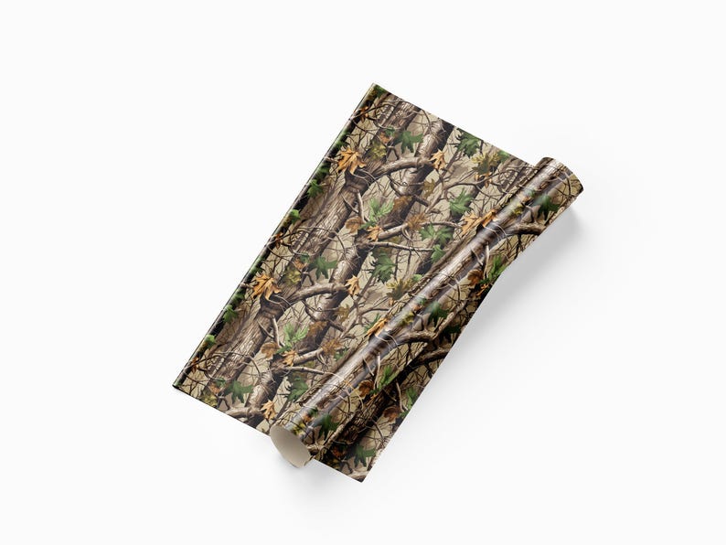 May include: A roll of camouflage pattern material. The pattern features a mix of brown, green, and tan colors, resembling a forest environment with branches and leaves. The material is partially rolled, revealing the pattern.