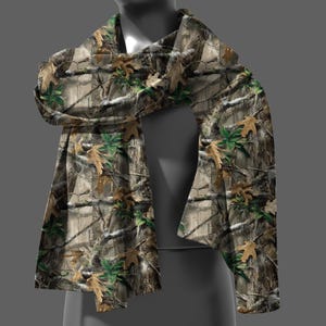 May include: A camouflage scarf with a pattern of tree branches, leaves, and bark in shades of brown, green, and tan. The scarf is draped over a mannequin, showcasing its design and texture.