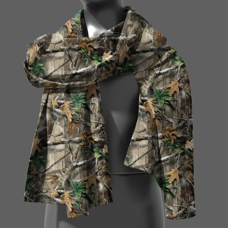 May include: A camouflage scarf with a pattern of tree branches, leaves, and bark in shades of brown, green, and tan. The scarf is draped over a mannequin, showcasing its design and texture.