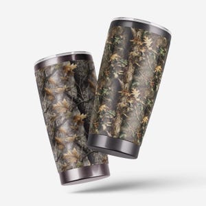 Puede incluir: Dos vasos aislados con un patr&oacute;n de camuflaje de hojas y ramas marrones, verdes y beiges. Los vasos tienen un borde y una base plateados, y un borde y una base negros. El dise&ntilde;o es un tema cl&aacute;sico al aire libre.