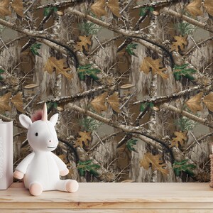 May include: A white unicorn plush toy with a pink horn and hooves sits on a wooden surface. Behind it is a camouflage pattern wallpaper with brown, green, and tan tones. Books and a small basket are also on the surface.