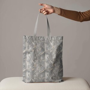 May include: A gray tote bag with a camouflage pattern of tree branches and leaves. The bag is held by a person's hand, showcasing its design and the length of the handles. The bag is made of a durable material.