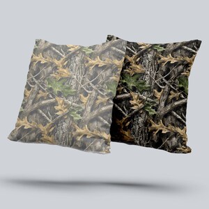 Puede incluir: Dos cojines cuadrados con un patr&oacute;n de camuflaje. El dise&ntilde;o presenta una mezcla de hojas y ramas de roble marrones, verdes y tostadas sobre un fondo moteado, creando un efecto de bosque naturalista.