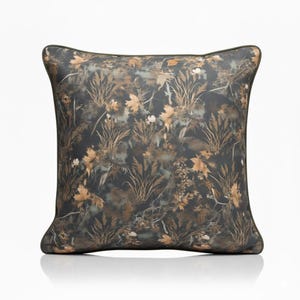 May include: Square decorative pillow with a nature-inspired pattern. The pillow features a dark background with a camouflage-like design of leaves, grasses, and other foliage in shades of brown, tan, and green. The pillow has a dark green border.