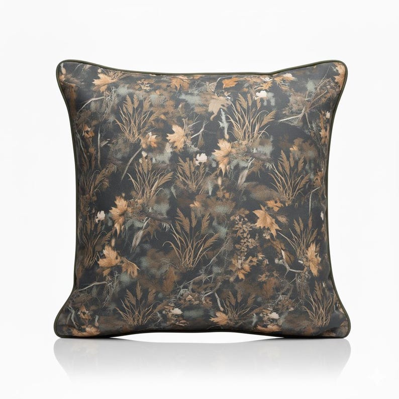 May include: Square decorative pillow with a nature-inspired pattern. The pillow features a dark background with a camouflage-like design of leaves, grasses, and other foliage in shades of brown, tan, and green. The pillow has a dark green border.