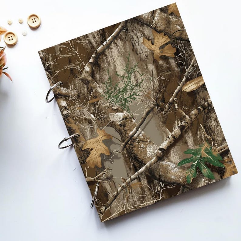 May include: A camouflage-patterned binder with three metal rings. The cover features a realistic design of tree branches, leaves, and bark in shades of brown, green, and tan. The binder is designed for organizing papers.