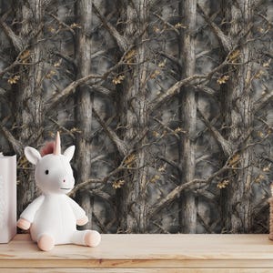 Puede incluir: Un juguete de peluche de unicornio blanco con cuerno y pezu&ntilde;as rosas est&aacute; sobre una superficie de madera. Detr&aacute;s, un papel pintado con un patr&oacute;n de camuflaje de troncos y ramas de &aacute;rboles. Tambi&eacute;n hay libros y una peque&ntilde;a cesta.