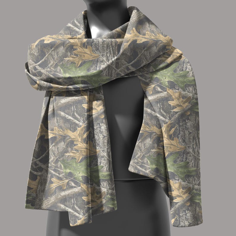 Puede incluir: Bufanda de camuflaje con un patr&oacute;n de hojas y ramas marrones, verdes y grises. La bufanda est&aacute; drapeada sobre un maniqu&iacute;, mostrando su dise&ntilde;o y textura. La tela parece suave y ligera.