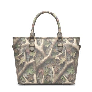 May include: A camouflage tote bag with a brown and green pattern featuring deer antlers and leaves. The bag has a top handle and metal hardware. The bag is designed with a shoulder strap.