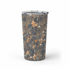 May include: A stainless steel tumbler with a clear lid. The tumbler features a camouflage pattern of autumn leaves, grass, and branches in shades of brown, tan, and gray. The design wraps around the entire body of the tumbler.