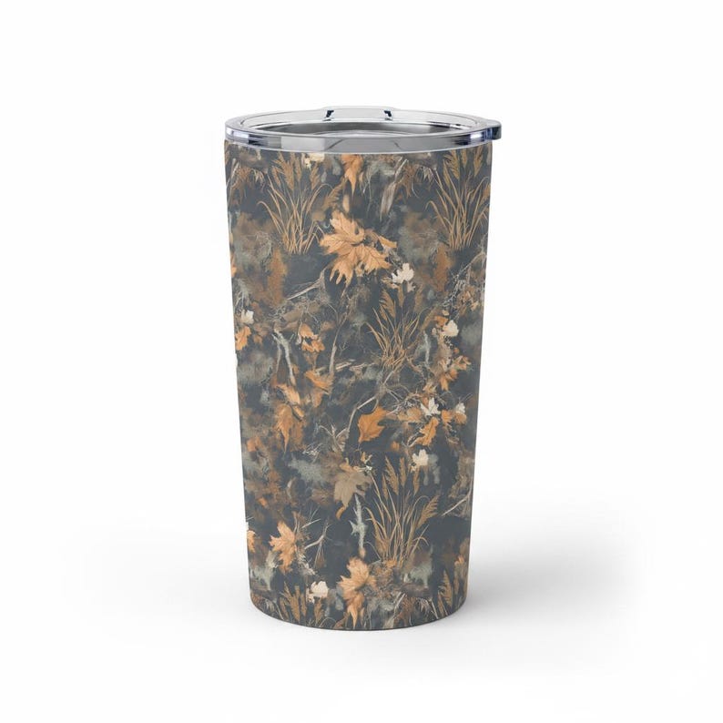 May include: A stainless steel tumbler with a clear lid. The tumbler features a camouflage pattern of autumn leaves, grass, and branches in shades of brown, tan, and gray. The design wraps around the entire body of the tumbler.