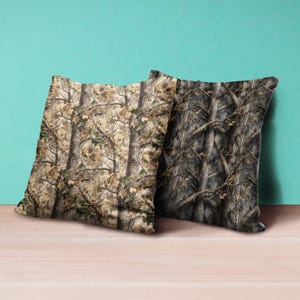Puede incluir: Dos cojines cuadrados con un patr&oacute;n de camuflaje. Un coj&iacute;n presenta un patr&oacute;n de &aacute;rboles marr&oacute;n claro y verde, mientras que el otro tiene un dise&ntilde;o de &aacute;rboles m&aacute;s oscuro, en tonos grises. Los cojines est&aacute;n colocados sobre una superficie de madera.