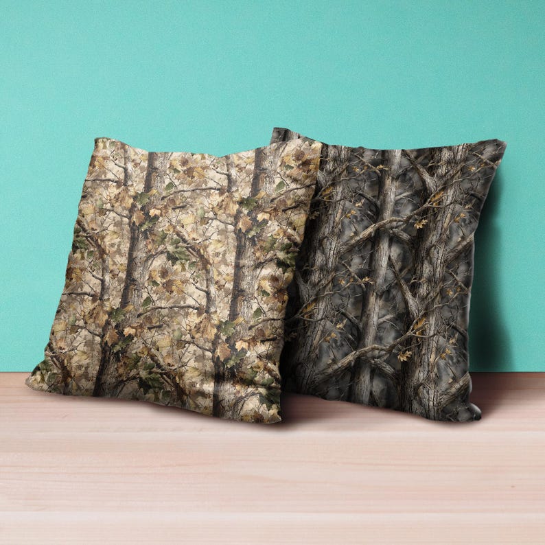 Puede incluir: Dos cojines cuadrados con un patr&oacute;n de camuflaje. Un coj&iacute;n presenta un patr&oacute;n de &aacute;rboles marr&oacute;n claro y verde, mientras que el otro tiene un dise&ntilde;o de &aacute;rboles m&aacute;s oscuro, en tonos grises. Los cojines est&aacute;n colocados sobre una superficie de madera.