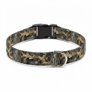 Puede incluir: Un collar para perros con un patr&oacute;n de camuflaje con hojas de roble y corteza de &aacute;rbol marrones y beige. El collar tiene una hebilla de pl&aacute;stico negro y una anilla en D de metal plateado para sujetar la correa.