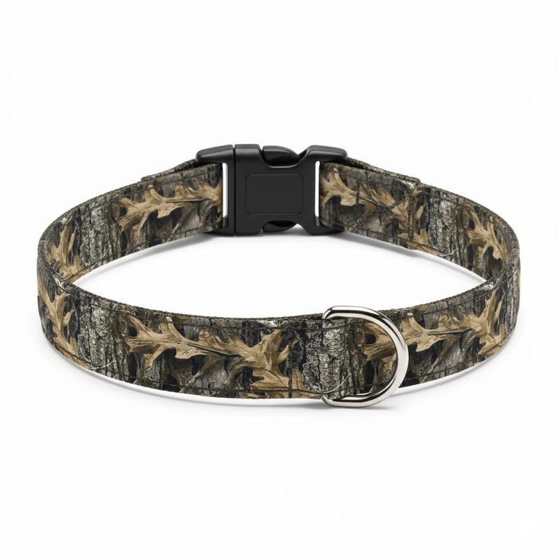 Puede incluir: Un collar para perros con un patr&oacute;n de camuflaje con hojas de roble y corteza de &aacute;rbol marrones y beige. El collar tiene una hebilla de pl&aacute;stico negro y una anilla en D de metal plateado para sujetar la correa.