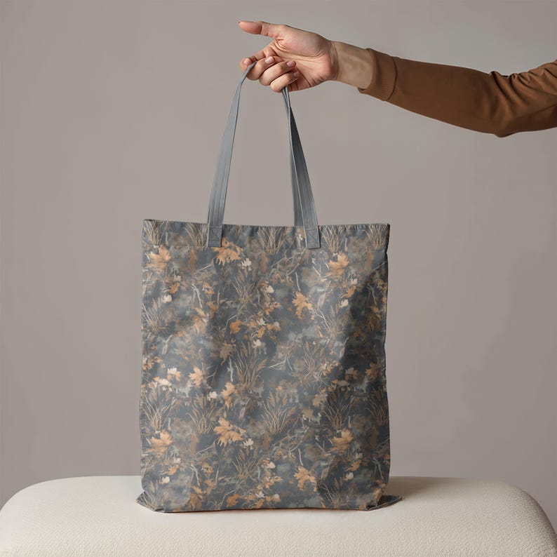 May include: A tote bag with a botanical pattern in shades of gray, brown, and beige. The bag has gray handles and is held up by a person's hand. The bag's design features a repeating pattern of leaves and foliage.