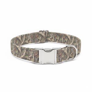 May include: A dog collar with a camouflage pattern in shades of brown, green, and tan. The collar has a silver metal buckle and a D-ring for leash attachment. The collar is designed for pets.