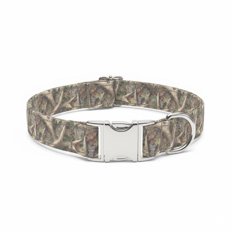 May include: A dog collar with a camouflage pattern in shades of brown, green, and tan. The collar has a silver metal buckle and a D-ring for leash attachment. The collar is designed for pets.