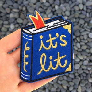 May include: A blue book patch with a red flame bookmark and the text "it's lit" in gold embroidery.
