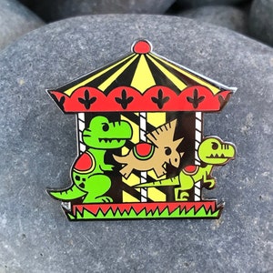 Op de afbeelding: Een emaille pin met een draaimolen met drie dinosaurussen. De draaimolen is rood, geel en zwart met groene, gele en bruine dinosaurussen.