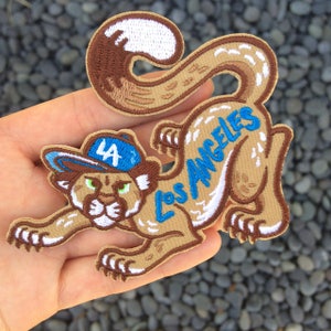May include: A brown embroidered patch of a cartoon cougar wearing a blue baseball cap with the letters "LA" on it. The cougar is jumping and the text "Los Angeles" is embroidered on its back.