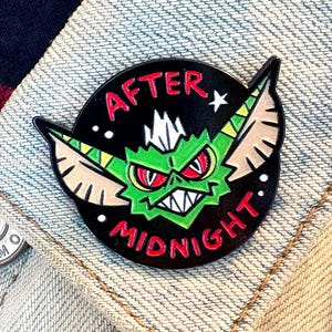 May include: A round black enamel pin with a green monster design and the words "AFTER MIDNIGHT" in red. The pin is attached to a light blue denim jacket. The jacket is over a red, white, and blue striped shirt.