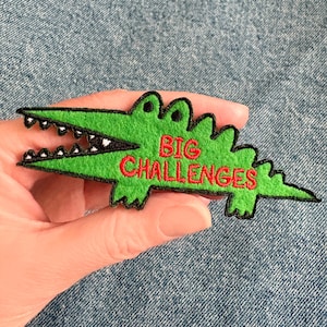 Peut inclure: Un écusson vert en forme d'alligator avec les mots "BIG CHALLENGES" brodés en rouge. L'écusson a un contour noir et des dents blanches. L'écusson est tenu dans une main.